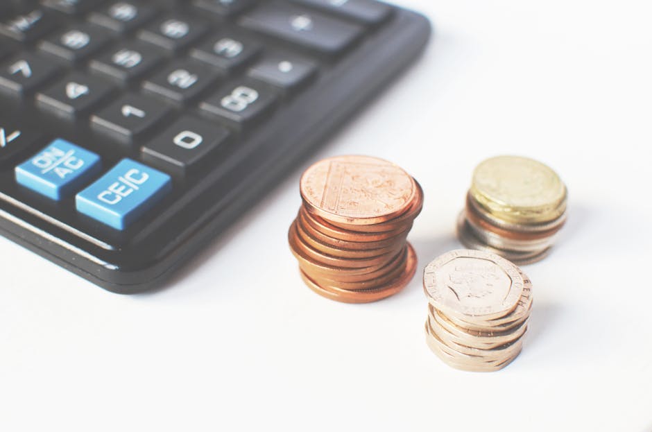 Close-up image showing a black calculator with blue and white buttons, placed next to three stacks of UK coins of various denominations on a white surface. The coins include copper-colored one-pence a